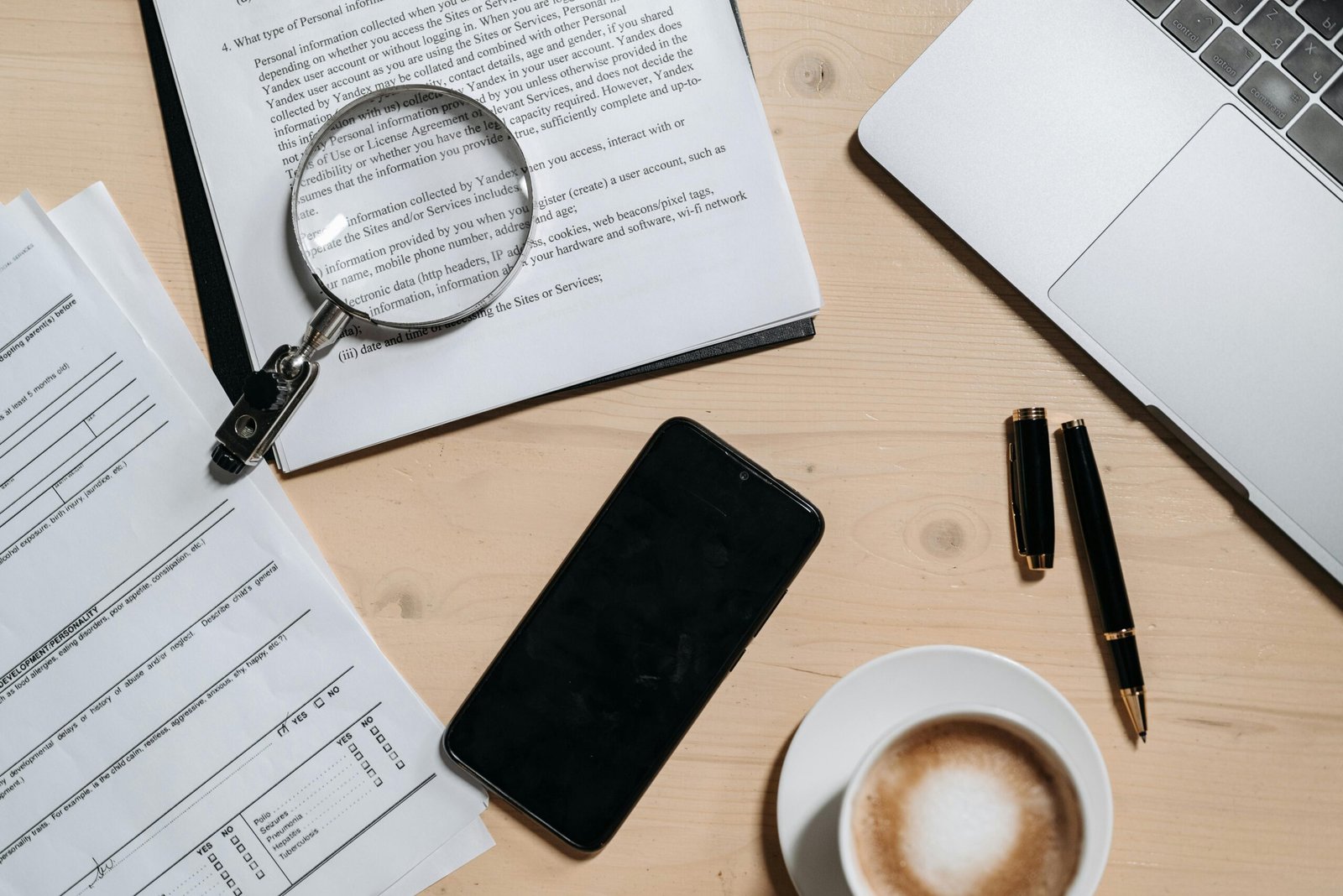 Overhead view of a desk with legal documents and forms, a magnifying glass, laptop, smartphone, pen, and a cup of coffee arranged on a wooden surface.